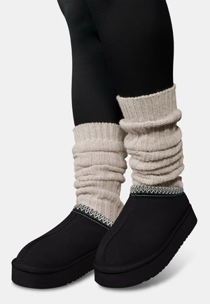 Black slip-on shoes with a thick sole and decorative stitching, paired with cream textured, ribbed leg warmers at the ankle.