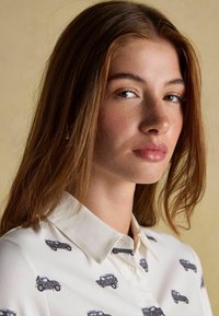 Biela košeľa s golierom s opakujúcim sa vzorom vintage áut v tmavomodrej a červenej farbe, zobrazená na neutrálnom pozadí.