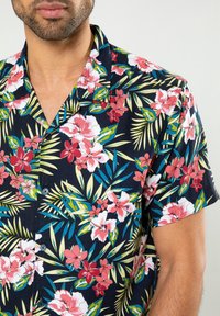 Hombre vestido con una camisa de manga corta negra con flores tropicales rosas y rojas y hojas verdes, mostrando el torso superior y el cuello.