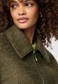 Vrouw met krullend haar, gekleed in een geweven olijfgroene ritsjas met een brede kraag, kijkt opzij tegen een neutrale achtergrond.