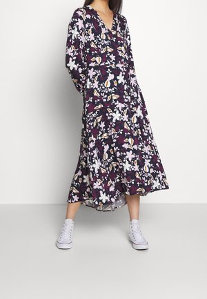 Robe florale en violet foncé ornée de fleurs blanches et roses, avec un décolleté en V, des manches longues et une longueur mi-mollet. Portée avec des baskets blanches.