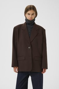 Blazer marron avec une fermeture à un seul bouton, deux poches avant et coupe décontractée. Porté par-dessus un haut gris foncé à col montant. Texture de tissu lisse.