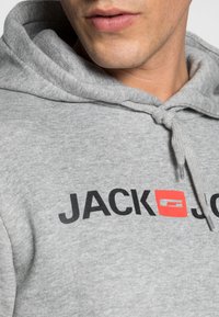Gray hoodie made of soft fabric. Features a kangaroo pocket, drawstring hood, and bold black text with a red accent reading "JACK & JONES."