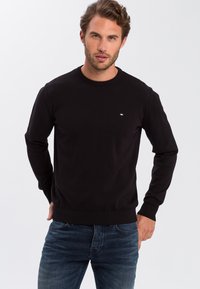 Czarny sweter dziany z długimi rękawami, okrągłym dekoltem i ściągaczem na dole. Mały detal z logo na piersi. W parze z teksturowanymi niebieskimi dżinsami.