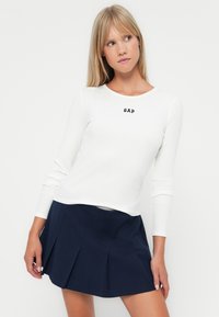 Wit geribd langärmelig top met een zwart "GAP" logo, gecombineerd met een marineblauwe geplooide rok. Beide stukken hebben een gladde textuur en klassieke ontwerpen.