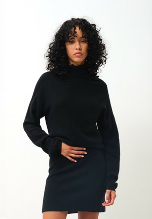 Musta neulepaita, jossa on korkea kaulus ja pitkät hihat, yhdistettynä saman sävyn mini-hameeseen; pehmeä rakenne ja slim fit -muotoilu.
