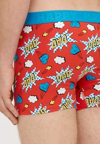 Sarkanas bokseršorti ar krāsainiem komiksu stila grafiskiem attēliem - mākoņiem, sirdīm un runas burbuļiem ar vārdu "TĒTIS", akcentēti ar zilu jostas daļu.