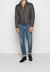 Vadderad svart jacka med hög krage, med en framdragkedja. Bärs över en beige topp, tillsammans med ljusblå denimjeans och svarta stövlar.