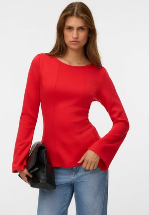 Femme portant un haut rouge moulant à manches longues et un jean bleu, tenant une grande pochette noire sur un fond uni.