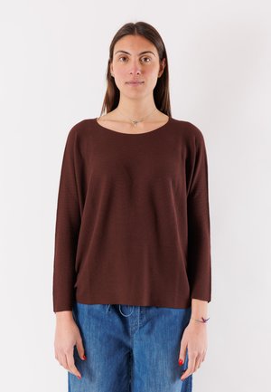 Donna con lunghi capelli castani che indossa una maglia marrone a maniche lunghe e jeans blu, in piedi dritta davanti a uno sfondo bianco semplice.