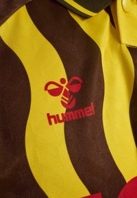 Brązowa i żółta koszulka polo w paski, wykonana z teksturowanej tkaniny, z czerwonym logo "hummel" oraz charakterystycznym kołnierzykiem.