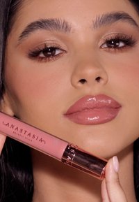 Płynna pomadka w przezroczystej tubce z metalową różowo-złotą nakrętką, przedstawiająca delikatny różowy odcień. Gładka konsystencja, nałożona na usta.