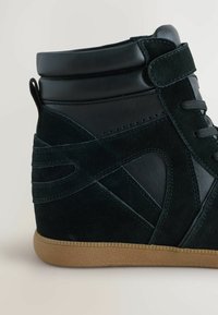 Sneaker alta in suede verde scuro e pelle nera. Presenta una suola in gomma testurizzata, caviglia imbottita e un frontale con lacci sicuri.