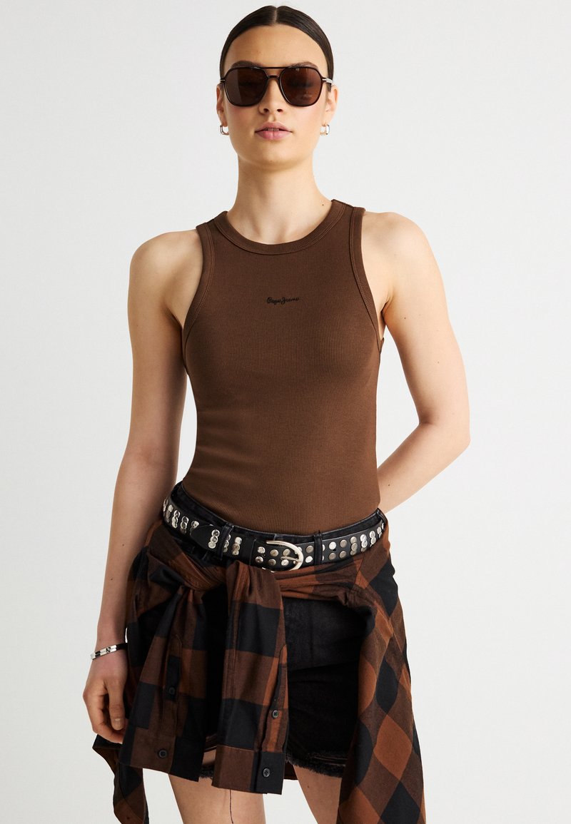 Γυναίκα που φοράει καφέ αμάνικο τοπ, μαύρα γυαλιά ηλίου, μαύρη ζώνη με καρφιά και δεμένο καφέ-μαύρο καρό πουκάμισο γύρω από τη μέση.