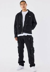 Chaqueta de mezclilla negra con cremallera frontal y múltiples bolsillos, combinada con pantalones negros estilo cargo que presentan cremalleras y bolsillos, usados con zapatillas blancas.