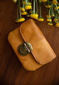 Rudos odos piniginė su apvalia forma, užsegama mosinėje sagtimi. Aplink kraštą matoma siūlų siuvimas, suteikiantis papildomą detalę.