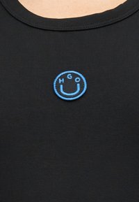 Μαύρο πουκάμισο με ένα μικρό μπλε κυκλικό σημάδι που απεικονίζει ένα χαμογελαστό πρόσωπο και τα γράμματα H, G, O κεντημένα μέσα στον κύκλο.