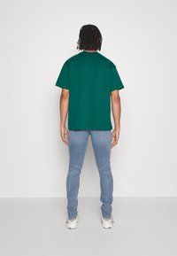 Zelené tričko s krátkým rukávem a volným střihem, pohled zezadu. V kombinaci se slim fit modrými džíny a bílými teniskami.