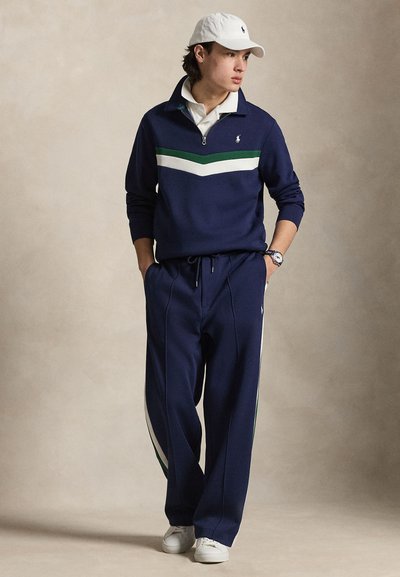 Jeune homme portant un survêtement bleu marine avec des bandes blanches et vertes, un polo blanc, une casquette blanche et des baskets blanches, debout les mains dans les poches.