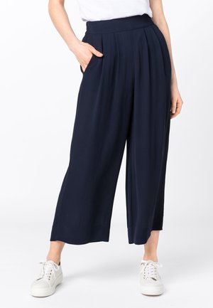 Vrouw draagt marineblauwe, wijde, cropped broek met plooien en zakken, gecombineerd met witte sneakers en een wit, ingestopt shirt.