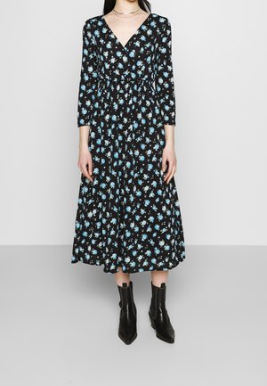 Femme portant une robe midi noire avec un imprimé floral bleu et blanc, des manches trois-quarts, un col en V et des bottines noires.