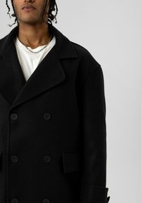 Persoon die een zwarte dubbelgeknoopte jas draagt over een wit shirt met een zilveren ketting, tegen een effen lichte achtergrond.