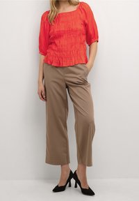 Blusa rossa arricciata con maniche a sbuffo, abbinata a pantaloni larghi color beige. Scarpe nere con tacco alto completano l'outfit.