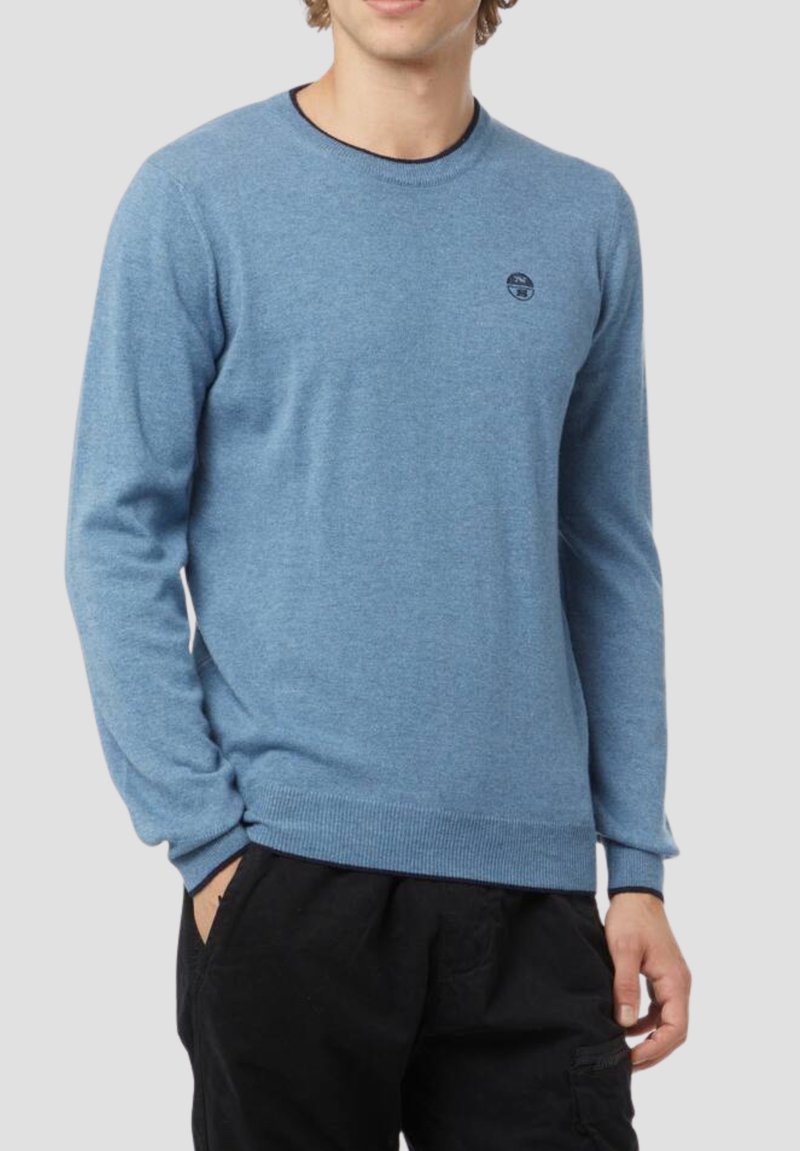 Giovane uomo che indossa un maglione girocollo azzurro chiaro con un piccolo logo rotondo sul petto e pantaloni neri con le mani nelle tasche.