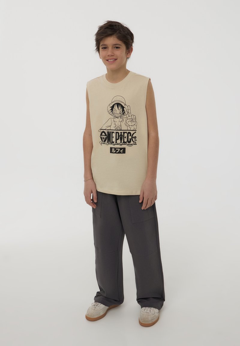 Ragazzo sorridente in piedi, con indosso una maglietta senza maniche beige di One Piece, pantaloni larghi grigi scuri e sneakers bianche su uno sfondo chiaro e semplice.