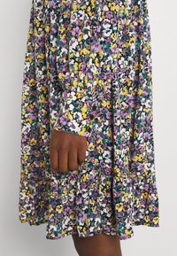 Fließendes Blumenkleid mit schwarzem Grundton, das mit weißen, gelben, lila und grünen Blumen verziert ist. Rüschensaum und Knopfdetails an den Ärmeln.
