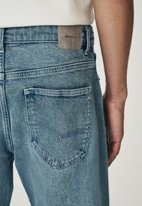 Blugi din denim de culoare bleu deschis, cu o croială clasică, prezintă o etichetă din piele la talie. Include două buzunare pe spate cu detalii subtile de cusătură.