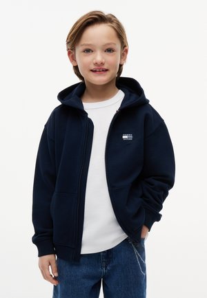Bambino piccolo con capelli biondi corti che indossa una felpa blu navy con zip sopra una t-shirt bianca e jeans blu, in piedi con una mano in tasca.