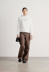 Hanorac alb cu glugă și logo gri, asortat cu pantaloni largi maro și încălțăminte neagră, ținând o geantă mică neagră.