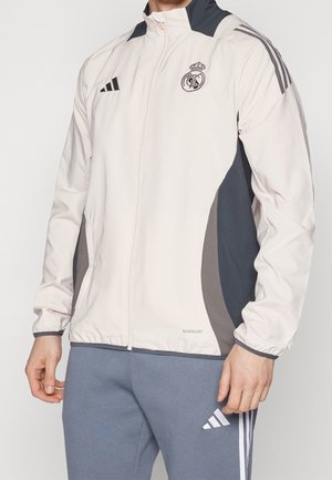 Man draagt een wit en donkergrijs Real Madrid voetbaljack met Adidas-logo's en grijze Adidas trainingsbroek.