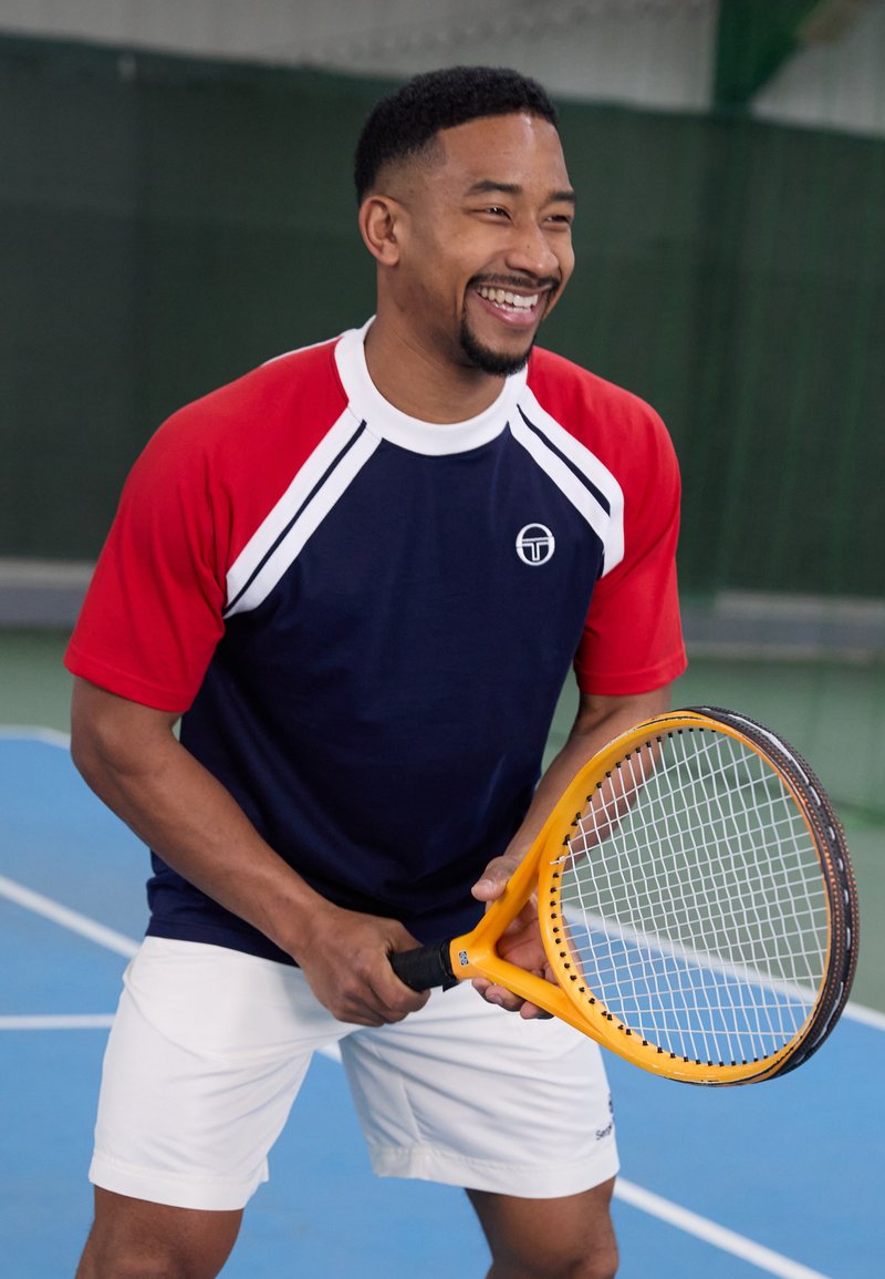 Campo da tennis blu con un uomo che indossa una maglia sportiva blu navy e rossa e pantaloni bianchi, mentre tiene una racchetta da tennis arancione con un'impugnatura gialla.