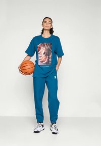 Tirkīza krāsas sporta komplekts ar īsām piedurknēm krekliņam, uz kura attēlota sneaker grafika, un atbilstošām sporta biksēm. Balti akcenti sneakeros.