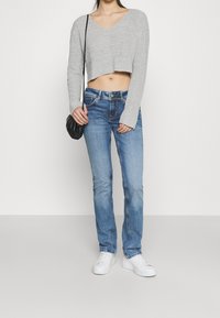 Maglione grigio a maniche lunghe, jeans dritti blu e sneakers bianche. Modella che tiene una piccola borsa nera.