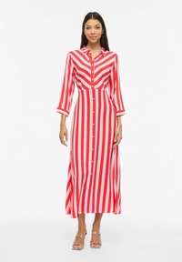 Femme portant une robe longue à rayures verticales rouges et blanches avec des boutons, des manches trois-quarts, et des talons ouverts argentés, debout sur un fond blanc.