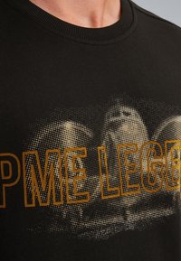 Camiseta de algodón negra con un estampado gráfico dorado con patrón de puntos y texto "PME LEGEND". Cuello acanalado, corte regular.