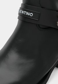 Botină din piele neagră, până la gleznă, cu o curea neagră ce prezintă logo-ul alb embossat "VALENTINO" și rivete argintii aproape de marginea de sus.