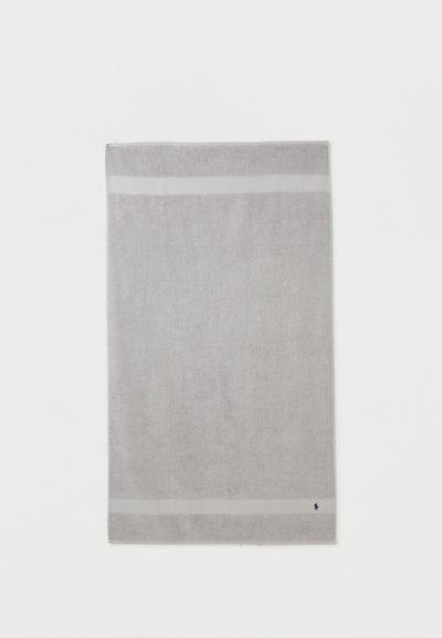 Ανοιχτό γκρι ορθογώνια πετσέτα με δύο οριζόντιες ανοιχτόχρωμες λωρίδες κοντά στις άκρες και μικρό σκούρο λογότυπο στην κάτω δεξιά γωνία.