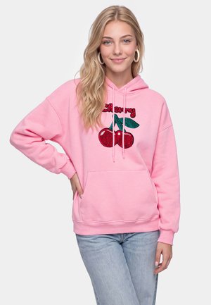 Ung kvinde med langt blondt hår iført store øreringe, en pink hoodie med røde kirsebær og lyseblå jeans, stående med den ene hånd på hoften.