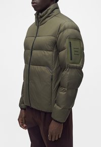 Homme portant une veste matelassée verte avec une fermeture éclair à l'avant et une poche zippée sur la manche gauche avec le logo "BOSS".