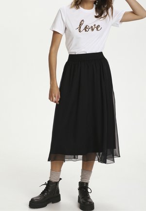 Kvinde iført hvid t-shirt med "love"-tryk, sort midinederdel, grå sokker og sorte snørebåndsstøvler, stående mod ensfarvet baggrund.