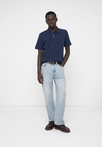 Ναυτικό μπλε polo μπλουζάκι με γιακά, ανοιχτό μπλε καμπάνες παντελόνια και καφέ παπούτσια. Απλός σχεδιασμός χωρίς ορατά μοτίβα ή λογότυπα.