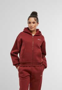 Bordová mikina na zips vyrobená z mäkkého materiálu, s prednou kapsou a rebrovanými manžetami, v kombinácii s nohavicami v rovnakom odtieni. Logo zobrazené vpredu.