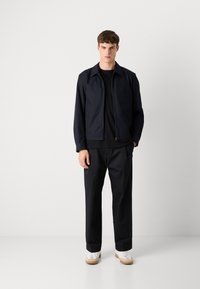 Casaco bomber azul-marinho com fecho éclair sobre uma camisola preta, combinado com calças pretas largas e ténis brancos com detalhes em castanho. Design minimalista.