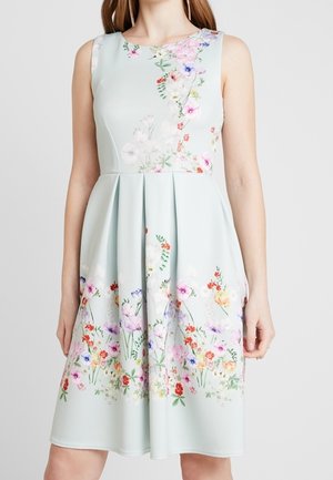 Bezrekawowa jasno-niebieska sukienka z kwiatowymi wzorami wokół talii i brzegu, z plisami i dopasowanym gorsetem na stojącej kobiecie.