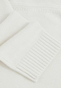 Detailný záber na bielu pletenú sveterovú rukáv, ktorý zobrazuje rebrový manžet a textúrovanú pleteninu.