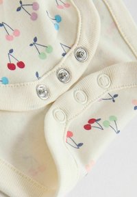 Vêtement pour bébé de couleur crème avec des boutons-pression et un motif de cerises multicolores imprimé sur le tissu.